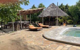 swimming pool with two sunbeds and covers terrace with chairs and round building with roof of palm leaves